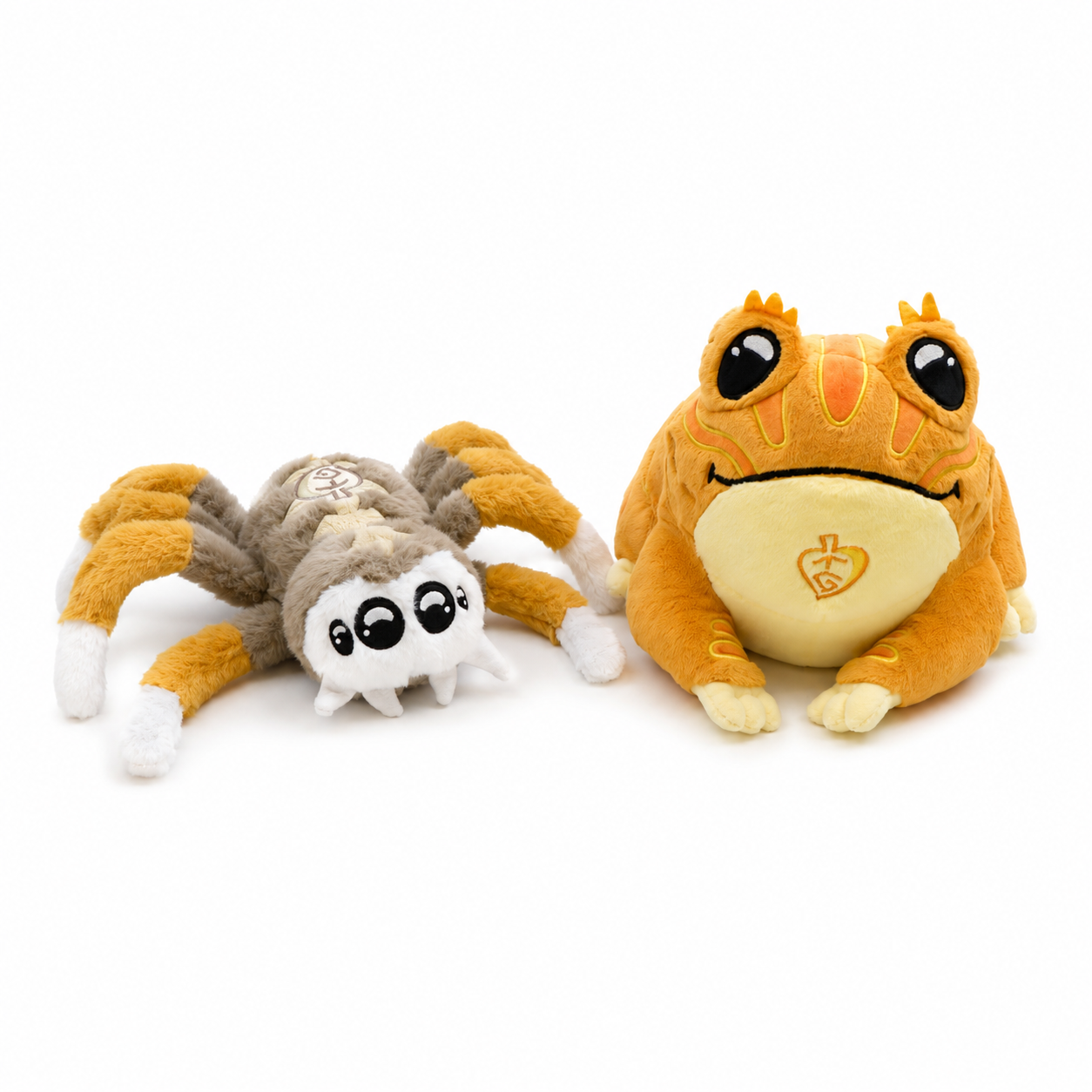 Two plush toys on a plain white background: a small spider plush with orange, brown, white, and gray coloring, and a larger frog plush in orange and pale yellow with a small heart emblem on its belly.